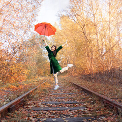young woman with orange umbrella jumping over the railroad tracks. Alley of autumn trees and...