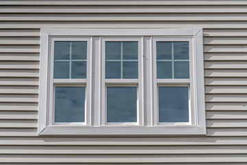 Triple hung window with fixed top sash and bottom sash that slides up, sash divided by white grilles a surrounded by white elegant frame  horizontal white vinyl siding on a new construction residence