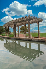 Long modern wooden pergola shade structure with reflection off the water fountain in a park with...