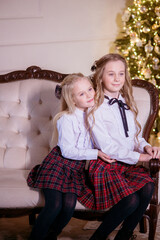 Lovely girls sisters with blonde curly hair with bans to the tols in classic school clothes by the Christmas tree in gold colors  in a classic interior. Christmas mood 
