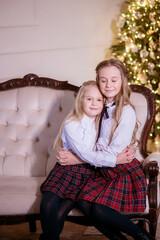 Lovely girls sisters with blonde curly hair with bans to the tols in classic school clothes by the Christmas tree in gold colors  in a classic interior. Christmas mood 
