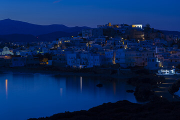Obraz premium Blue night over a mountainous Greek island with a white city. Street lights are reflected in the calm sea