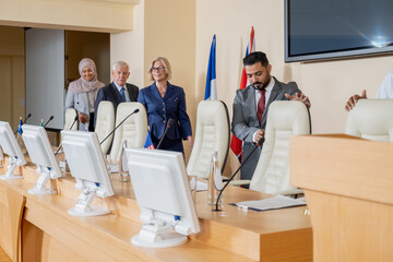 Group of multi-ethnic participants coming into conference room and taking seats in front of monitors