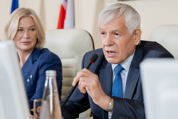 Senior Caucasian politician in formal suit sitting at conference table and using microphone while...