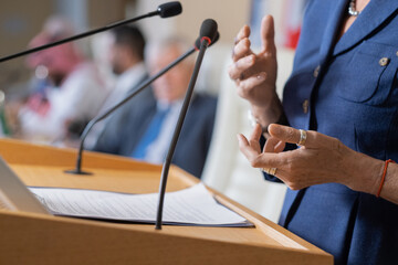 Close-up of unrecognizable female politician in rings standing at rostrum with microphones and clipboard while addressing conference - obrazy, fototapety, plakaty