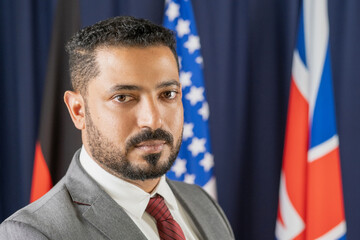 Portrait of serious mixed race bearded politician in suit posing against national flags in congress hall - obrazy, fototapety, plakaty