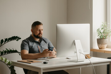 A smiling man works remotely on a desktop computer. A guy with a beard is holding a cellphone during a report of a colleague at a video conference at home. A teacher is preparing for an online lecture