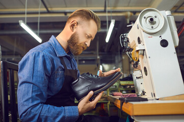 Adult caucasian man shoemaker in uniform holding handmade leather boot checking for defects. Craftsman in apron working at workplace. Shoe factory, footwear industry and cobbler shop concept