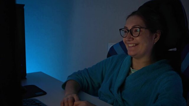 Brunette woman in eyeglasses and blue bathrobe watches TV series with bright smile and interest on large computer screen sitting in dark room.