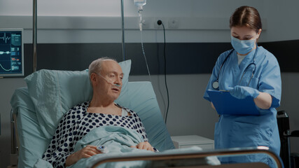 Hospital ward nurse checking on sick patient in bed at medical recovery facility. Old man with modern oximeter and nasal oxygen tube getting help for treating healthcare problems
