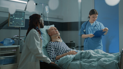African american doctor talking to sick patient in hospital ward and nurse checking symptoms. Old man with cervical neck collar sitting in bed at intensive care room to heal problems