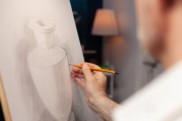 Close up of caucasian hand of old man holding pencil on canvas, drawing vase model at art studio. Senior person working as artist creating authentic masterpiece in workshop space