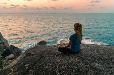 Fototapeta na wymiar Young woman in lotus position meditates on rocks by the sea at sunrise. Colorful sky on the background.
