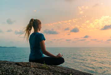 Young woman in lotus position meditates on rocks by the sea at sunrise. Colorful sky on the background.