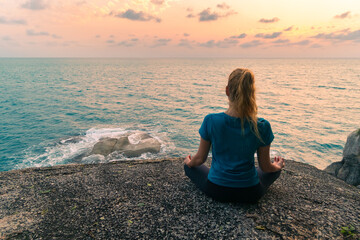 Young woman in lotus position meditates on rocks by the sea at sunrise. Colorful sky on the...
