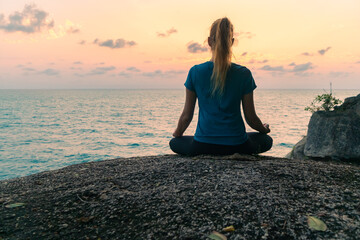 Young woman in lotus position meditates on rocks by the sea at sunrise. Colorful sky on the...