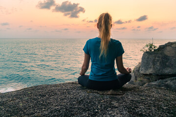 Fototapeta na wymiar Young woman in lotus position meditates on rocks by the sea at sunrise. Colorful sky on the background.