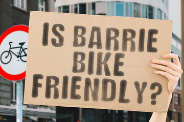 The question " Is Barrie bike friendly? " on a banner in men's hand with blurred background. Transportation. Zero waste. Bicycle lane. Streets. City. Safety. Insecure. Road signs. Dangerous