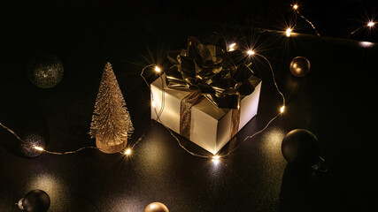 Holiday Christmas background. White gift box with golden ribbon, New Year balls and sparkling lights garland in xmas composition on black for greeting card. Winter holidays, New Year.