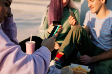 Happy teenage friends are sitting in urban exterior and using their phones for hanging on social...