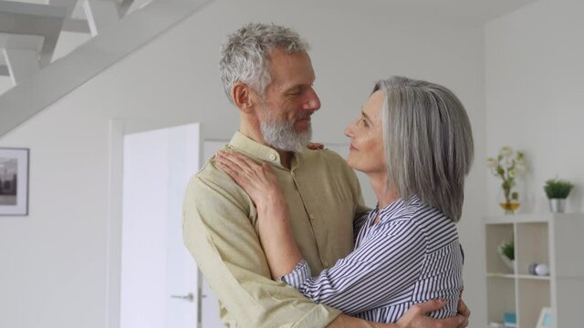 Dreamy senior adult mature couple standing hugging, bonding, thinking of good future. Carefree cheerful mid age old husband hugging wife looking away dreaming, enjoying wellbeing love in new house.