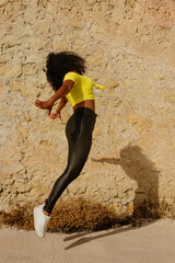 Latin black girl jumps for joy outdoors, yellow wall background