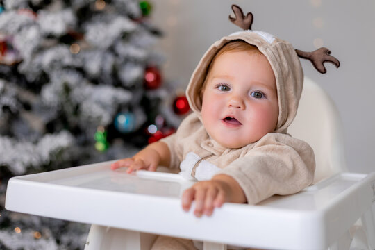 portrait of chubby cheeked baby in a jumpsuit with deer horns sits in a white high chair for feeding on the background of Christmas tree. winter New Year's concept. space for text. High quality photo
