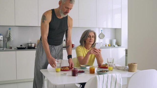 Happy older mature family couple after breakfast, using mobile apps on cell phone in kitchen, man cleaning table. Smiling senior husband and wife talking, looking at phone enjoying morning together.