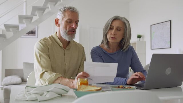 Senior middle aged couple holding financial documents paper bills paying bank loan online, calculating pension fees, payments, taxes, planning family retirement money finances using laptop at home.
