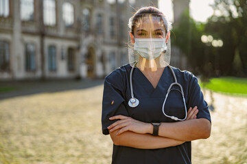 Naklejka premium Cheerful young woman nurse wearing face shield and mask for safety to prevent Covid19, looking at camera, standing outdoors on a sunny day