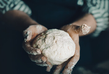 A male chef in a dark blue apron holds a pastry dough in his hands. Home cooking. Traditional pizza recipe. Delicious food.