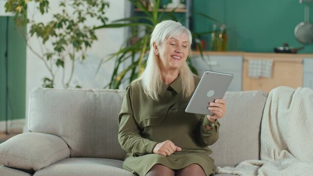 A Happy Elderly Woman Makes an Online Call Using Her Tablet, Enjoys Online Chatting with Her Family While Sitting on the Couch at Home. Elderly People Modern Technology and Communication Concept.