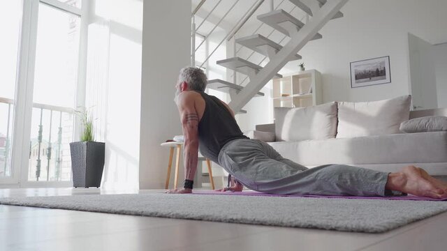 Fit sporty active tattooed senior mature mid age hipster man doing morning workout exercise at home, enjoying sport pilates yoga fitness training on mat in modern living room.