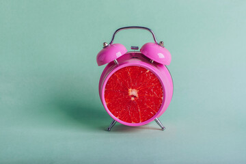 Pink alarm clock with grapefruit inside isolated on green background in a healthy morning wake up...
