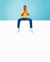 Fototapeta na wymiar Cool black man sitting on blank white banner, touching his face, shouting OMG in excitement, mockup for your ad design