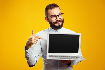 Cheerful hipster man with beard wearing beige shirt and eyeglasses pointing at new laptop in hands...