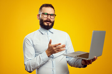 Young bearded man holding laptop in hands while looking at camera with confusion, isolated over yellow background