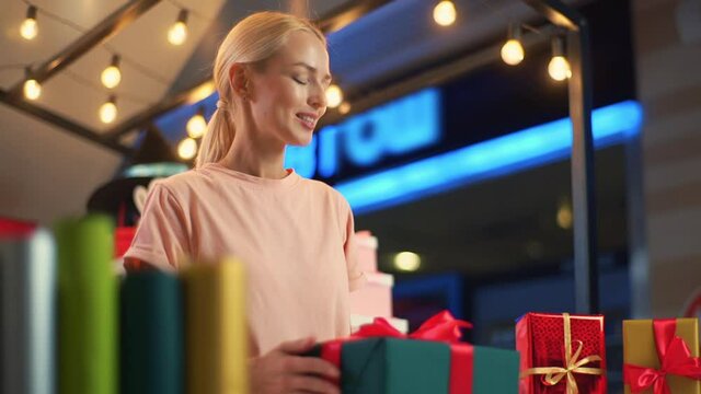 Low-angle view of young cheerful woman seller wrapping Christmas gift box tying red ribbon and decorating beautiful bow standing at table of holiday store, blurred background of bright lighting.
