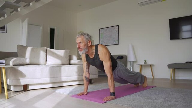 Fit healthy sporty active tattooed senior mature mid age hipster man doing morning workout exercise at home, enjoying sport pilates yoga fitness training breathing on mat in modern living room.