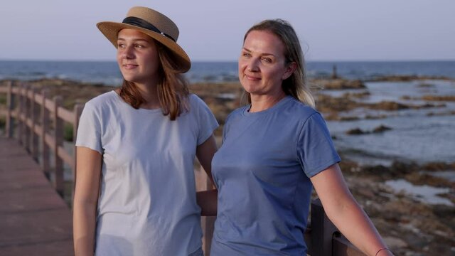 Mother and daughter standing on wooden sidewalk by the sea at sunset and talking. A middle-aged woman and teenage girl standing side by side and wearing blue t-shirts and shorts. Motherhood concept