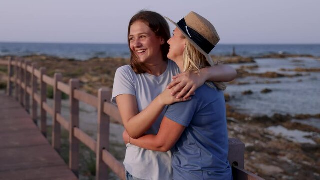 Mother and Daughter embracing on wooden sidewalk by the sea at sunset. A middle-aged woman and teenage girl standing side by side and wearing blue t-shirts and shorts. Motherhood and travel concept