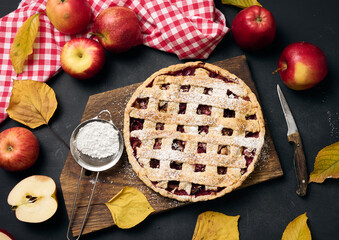 baked round traditional apple pie on brown wooden board and fresh red apples, top view