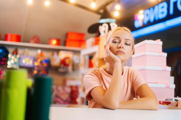 Close-up of sad attractive young sales woman sitting at counter of holiday store in mall waiting for customers. Small business suffers during Christmas sales because of lockdown caused by coronavirus.