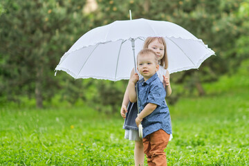 little girl and boy are hiding under an umbrella. brother and sister are playing in the park.