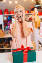 Vertical portrait of excited cheerful young woman standing by counter table with wrapped Christmas gift box tied red ribbon and decorated beautiful bow, bright blurred background of festive packages.