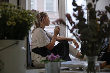 Artist Woman in dirty apron resting with a cup of tea sitting on the windowsill and looks out the window