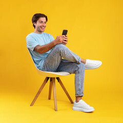 Happy guy using mobile phone at studio, sitting on chair - obrazy, fototapety, plakaty