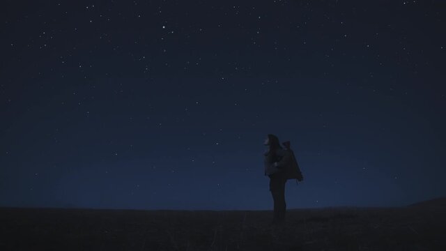 The woman with camera bag standing against the starry sky with falling stars in the middle of a field at night. Woman looking up to the stars. Zoom out. 