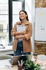 Happy moment. Brunette woman holding folder with documents and smiling with closed eyes at her loft office. Stock photo