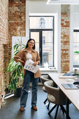 Full length view of the confident businesswoman wearing casual clothes holding documents and diagrams and smiling to the camera at her loft office